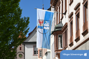 An einer Hausfassade hängt eine blau-weiß gestreifte Fahne mit Stadtwappen: roter Adler, weißer Schwan und Wellen, vor blauem Himmel.