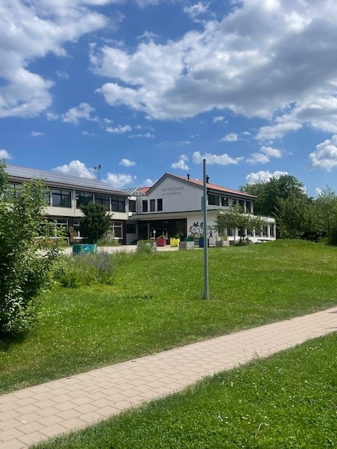 Zweistöckiges öffentliches Gebäude mit weißer Fassade und rotem Dach, davor großer grüner Rasen und gepflasterter Weg, blauer Himmel mit weißen Wolken.