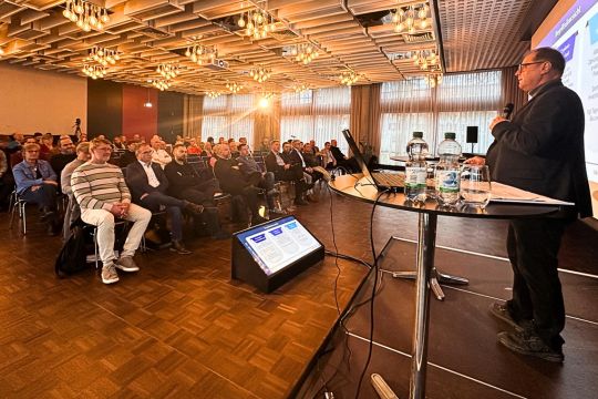 Oberbürgermeister Jürgen Roth steht am Podium und hält eine Präsentation vor einem großen Publikum in einem Konferenzraum; Zuhörer sitzen in Reihen, auf einer Leinwand sind Folien zu sehen.