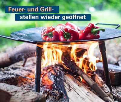 Rote Paprika rösten auf einer Grillpfanne über offenem Lagerfeuer. Bildtext: „Feuer- und Grillstellen wieder geöffnet“.