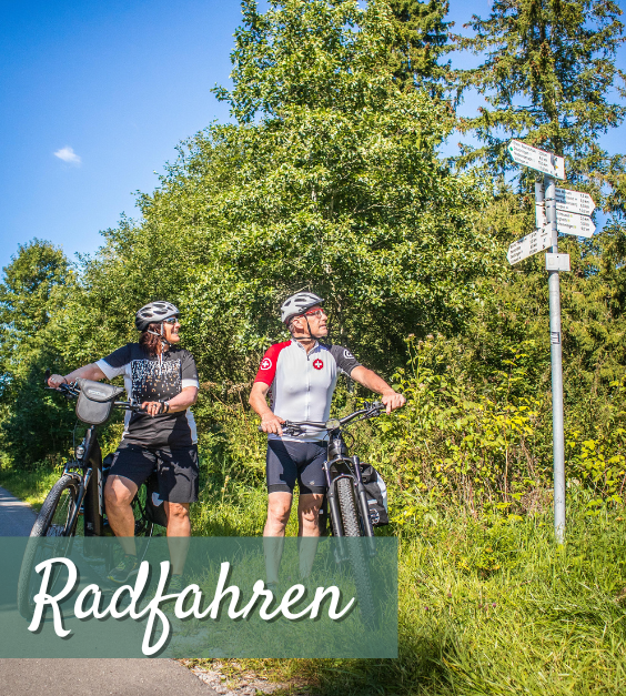 Zwei Radfahrer mit Helmen stehen neben ihren Fahrrädern auf einem Waldweg und schauen auf einen Wegweiser.