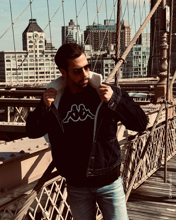 Ein Mann mit Sonnenbrille steht auf einer Brücke, trägt eine Jeansjacke und ein T-Shirt mit Logo.