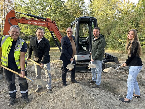 Fünf lächelnde Personen mit Schaufeln vor einem Bagger, die symbolisch Erde aus einem Erdhaufen heben bei einem Spatenstich auf einer Baustelle.