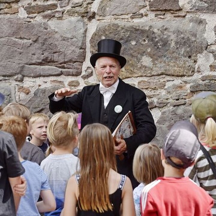 Ein Mann in einem schwarzen Anzug und Zylinder erzählt einer Gruppe von Kindern eine Geschichte. (KI generiert)