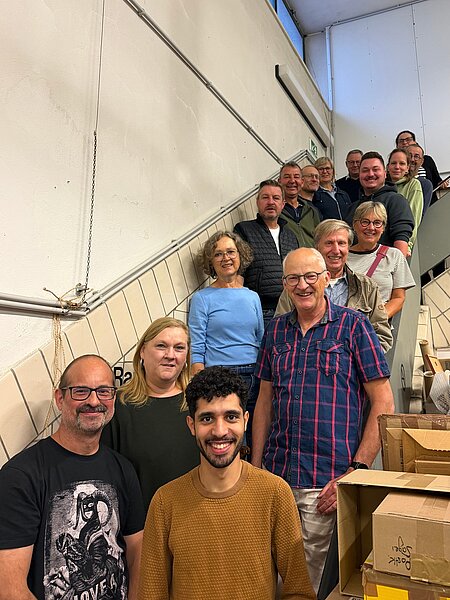 Etwa 15 lächelnde Personen unterschiedlichen Alters in einer Treppenhausreihe, frontal zur Kamera aufgestellt; Kartons stehen neben der Treppe.