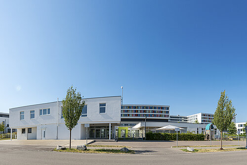 Moderner weißer Schul- oder Bürokomplex mit mehreren Gebäudeflügeln, Eingangsbereich, Sitzecken mit Sonnenschirmen, jungen Bäumen und klarem blauem Himmel.