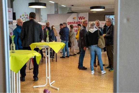 Mehrere Menschen stehen in einem hellen Veranstaltungsraum und unterhalten sich an Stehtischen mit gelben Tischdecken; im Hintergrund Informationswände und ein Banner mit der Aufschrift „Wir gestalten“ – Szene einer öffentlichen Bürger- oder Informationsveranstaltung.
