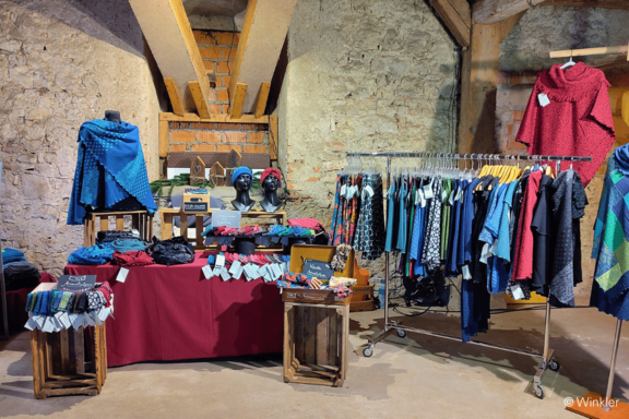 Marktstand in einem rustikalen Raum mit bunten Strickkleidern und Ponchos an einem Kleiderständer, Tischen mit Schals, Mützen, Accessoires und zwei Schaufensterbüsten.