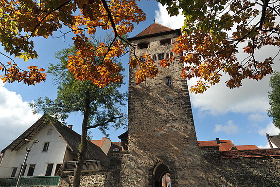 Mittelalterlicher steinerner Stadtturm mit Torbogen und Zinnen, flankiert von alten Häusern; im Vordergrund herbstlich orange gefärbte Blätter, im Hintergrund blauer Himmel mit Wolken.