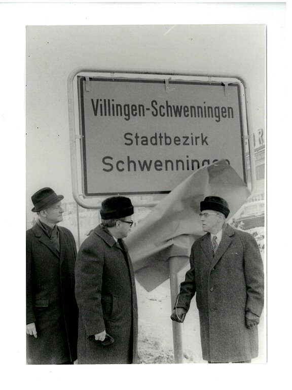 Drei Männer in Mänteln und Hüten enthüllen ein Ortsschild mit der Aufschrift „Villingen‑Schwenningen Stadtbezirk Schwenningen“.