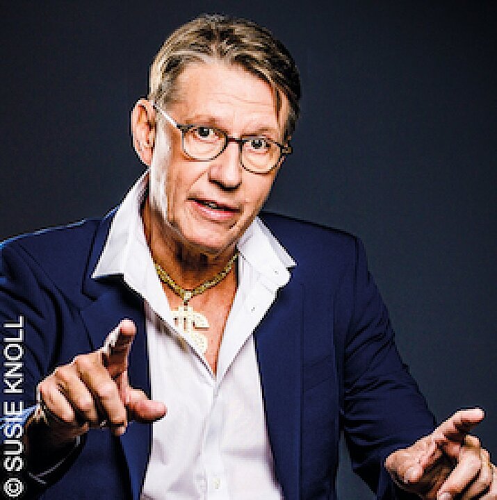 Ältere Person mit Brille, grauen Locken und blauem Blazer hält Mikrofon und lächelt. (KI generiert)