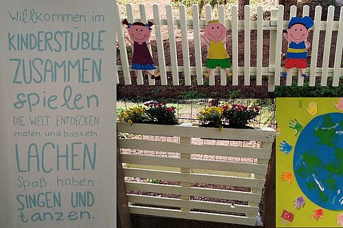 Willkommensschild "Kinderstüble" mit Text über gemeinsames Spielen, Entdecken, Lachen, Singen und Tanzen; davor ein weißer Lattenzaun mit bunten Papiersilhouetten von Kindern und ein Plakat der Erde, umgeben von bunten Handabdrücken.