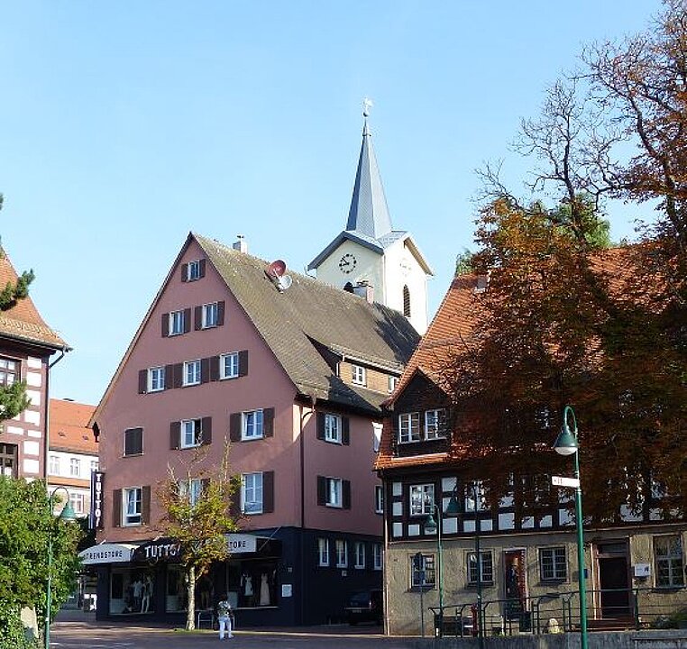 Ein Gebäude mit einem spitzen Turm und einer Uhr, umgeben von Bäumen und anderen Gebäuden. (KI generiert)