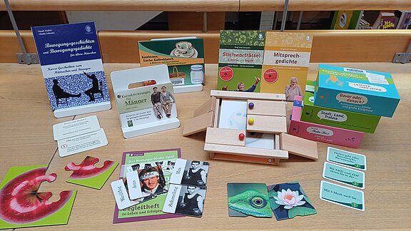 Auslage verschiedener kognitiver Stimulationsspiele und -materialien auf einem Tisch: ein Holzspiel mit bunten Kugeln, Bild- und Fragekarten, kleine Boxen und Broschüren.