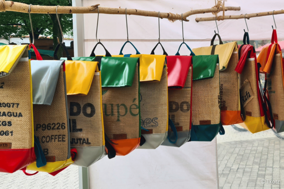 Recycelte Handtaschen aus Jute-Kaffeesäcken mit bunten Kunststoff-Applikationen, nebeneinander an einem Holzast aufgehängt, als Marktverkauf präsentiert.
