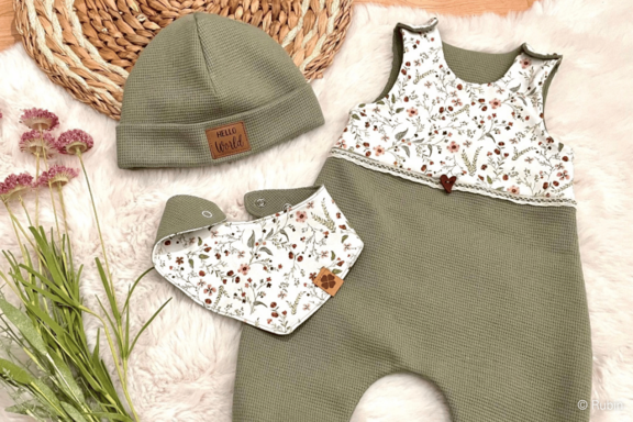 Babyset: olivgrüner, ärmelloser Strampler mit floralem Oberteil und Herzknopf, dazu passende olivgrüne Mütze und geblümtes Halstuch/Lätzchen, auf weichem Untergrund arrangiert.