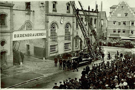 Schwarzweiß-Foto: Große Menschenmenge und Feuerwehr mit ausgefahrener Leiter vor dem Tor der „Bärenbrauerei“.