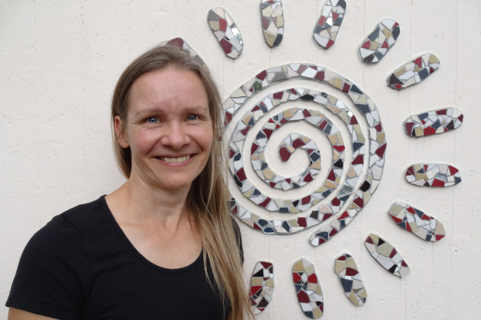 Lächelnde Frau mit langem Haar und schwarzem T‑Shirt steht vor einer weißen Wand neben einer Spiral‑Mosaiksonne.