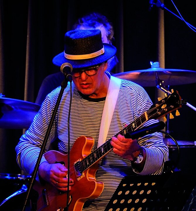 Sänger mit Gitarre auf der Bühne, trägt gestreiftes Hemd und Hut. (KI generiert)