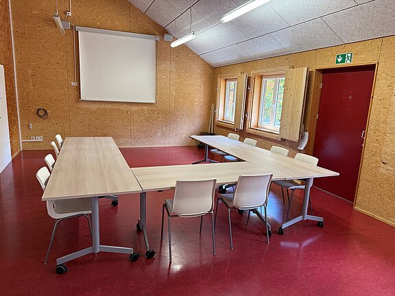 Leerer Besprechungsraum mit U-förmig aufgestellten Tischen und weißen Stühlen, Leinwand an der Wand, drei Fenster, rote Tür sowie helle Holzverkleidung und roter Boden.