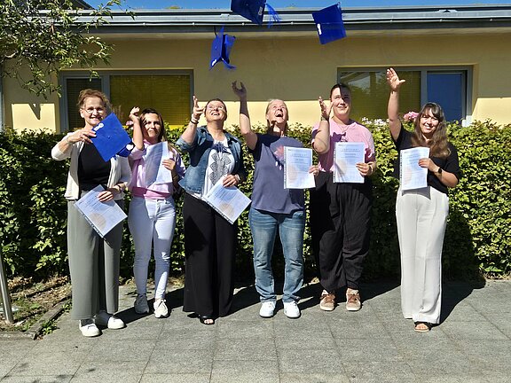 Sieben Personen stehen nebeneinander vor einem Gebäude, halten Zertifikate und werfen blaue Hüte in die Luft, feiern sichtbar ihren Abschluss.