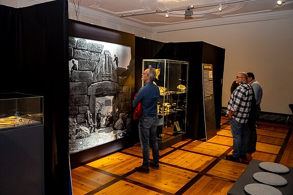 In einem Museum stehen mehrere Besucher vor einer Wand mit einer historischen schwarz-weißen Fotografie, die Arbeiten an einem alten Steinbau zeigt. In Vitrinen sind verschiedene archäologische Artefakte ausgestellt. Die Umgebung wirkt dunkel, um die Exponate hervorzuheben.
