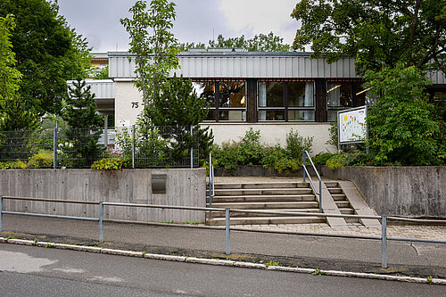 Eingangsbereich eines eingeschossigen Gebäudes mit Hausnummer 75: breite Treppe und Rampe mit Handläufen, niedrige Betonmauer, Vorgarten mit Sträuchern und Bäumen, große Fenster und ein Schild mit bunter Kinderzeichnung – wirkt wie Kindergarten oder Schule.