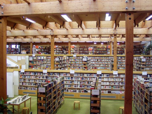 Großer, heller Bibliotheksraum mit sichtbaren Holzbalken, mehreren Reihen voller Bücherregale auf zwei Ebenen und vereinzelten Sitzplätzen.