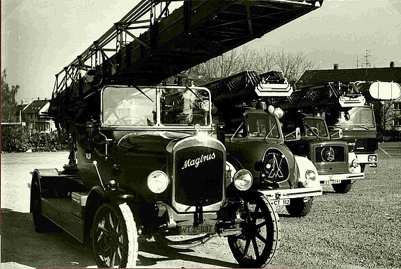 Vier historische Feuerwehrfahrzeuge in Reih und Glied auf einem Hof, vorne ein alter Magirus-Leiterwagen mit ausgefahrener Drehleiter, dahinter zwei weitere klassische Löschfahrzeuge und ein modernerer Drehleiterwagen.