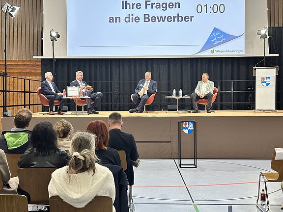 Podiumsdiskussion in einer Halle: vier sitzende Gesprächsteilnehmer auf der Bühne, Publikum im Vordergrund, große Projektionswand mit dem Text „Ihre Fragen an die Bewerber“ und eingeblendetem Timer.
