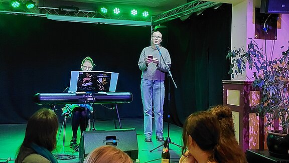 Auf einer Bühne steht eine Person am Mikrofon und liest vor; links begleitet eine Person das Geschehen am Keyboard. Publikum im Vordergrund.