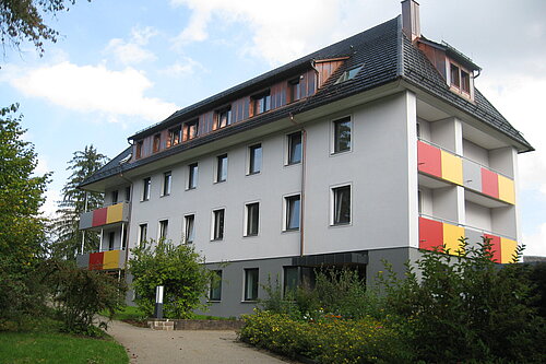 Mehrstöckiges Wohnhaus mit weißer Fassade, dunklem Satteldach und Balkonen mit gelb-roten Brüstungen; Vorgarten und Gehweg führen zum Eingang.