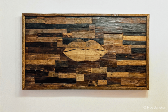 Rechteckiges Wandbild aus unterschiedlich gefärbten Holzplanken in Mosaikmuster; mittig ein helleres, stilisiertes Lippenmotiv, eingefasst in Holzrahmen und an einer weißen Wand aufgehängt.