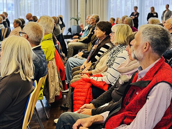 Älteres Publikum sitzt in Reihen in einem Konferenzraum und hört aufmerksam einer Veranstaltung oder einem Vortrag zu.
