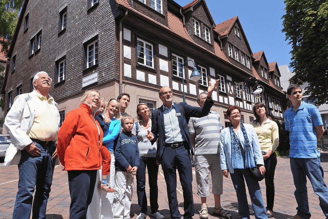 Gruppe von Menschen vor einem Fachwerkhaus, einige zeigen auf ein Schild. (KI generiert)
