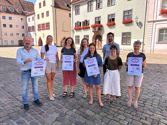 Acht Personen stehen in einer Reihe auf einem gepflasterten Marktplatz vor historischen Häusern; fünf halten Informationsplakate und alle blicken lächelnd zur Kamera.