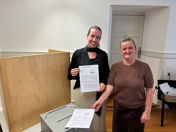 Zwei lächelnde Frauen in einem Innenraum: eine hält ein Dokument hoch, die andere schiebt ein Formular durch den Schlitz einer grauen Box. Im Hintergrund Trennwand und Tür.