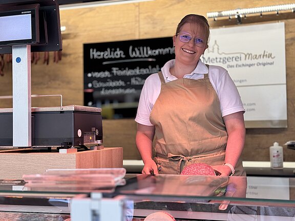 Lächelnde Verkäuferin in Schürze steht hinter einer Metzger-Theke, hält Wurstplatte; im Vordergrund Glasscheibe und Kassenbereich, freundliche Marktszene.