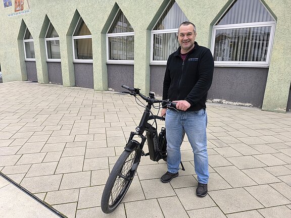 Ein lächelnder Mann steht auf einem gepflasterten Platz und hält ein schwarzes Elektrofahrrad vor einem Gebäude mit dreieckigen Fenstern.