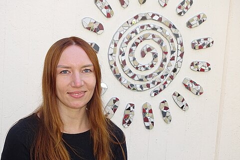 Person mit langen roten Haaren und schwarzem Oberteil lächelt vor einer weißen Wand mit einer spiralförmigen Mosaik-Sonne.