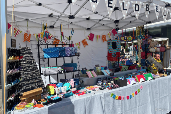 Marktstand unter einem weißen Pavillon mit bunten, handgemachten Accessoires: Geldbörsen, Täschchen, Schmuck, Schlüsselanhänger und kleine Geschenkartikel, auf Tischen und Regalen arrangiert, dekoriert mit bunten Wimpelgirlanden.