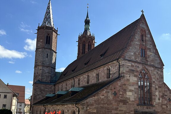Blick auf eine historische Sandsteinkirche mit zwei Türmen; rote Blumen im Vordergrund und blauer Himmel.