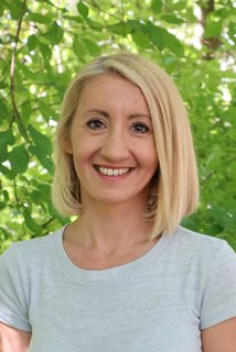 Porträt einer lächelnden Frau mit schulterlangen blonden Haaren und hellgrauem T‑Shirt, stehend vor dichtem grünem Laub.