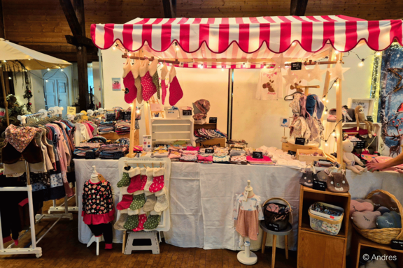 Marktstand mit rot-weiß gestreifter Markise, gefüllt mit Babykleidung, Kinderkleidern, Socken, Schuhen und Accessoires auf Tischen, Regalen und Puppenständern.