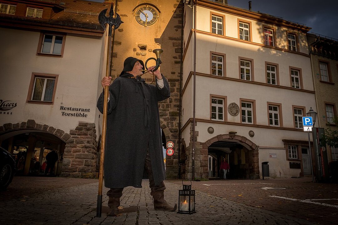 Eine Person in historischer Kleidung steht auf einem Kopfsteinpflasterplatz und bläst in ein Horn. Im Hintergrund sind Gebäude mit Uhren und ein Restaurant. (KI generiert)