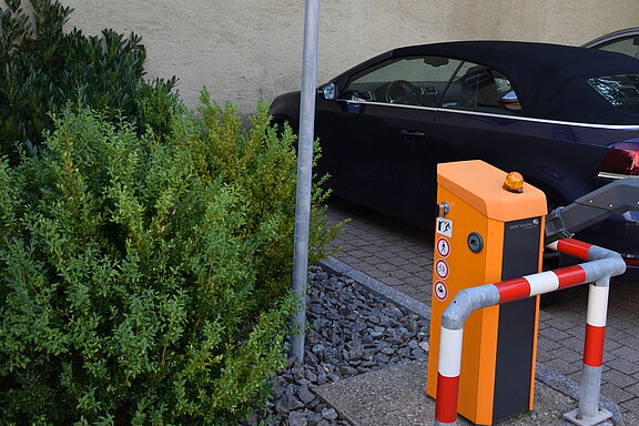 Privatparkplatz: Informationsschild an einem Mast, orangefarbene Schrankenanlage mit Parkkassette und Absperrpfosten, ein geparktes dunkles Auto und Büsche vor einer hellen Hauswand.