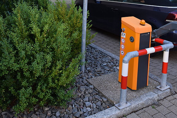 Privatparkplatz: Informationsschild an einem Mast, orangefarbene Schrankenanlage mit Parkkassette und Absperrpfosten, ein geparktes dunkles Auto und Büsche vor einer hellen Hauswand.