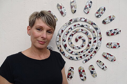 Person mit kurzem Haar in schwarzem T‑Shirt steht lächelnd vor einer weißen Wand, daneben ein dekoratives Mosaik in Form einer Spirale/Sonne aus roten, grauen und weißen Scherben.