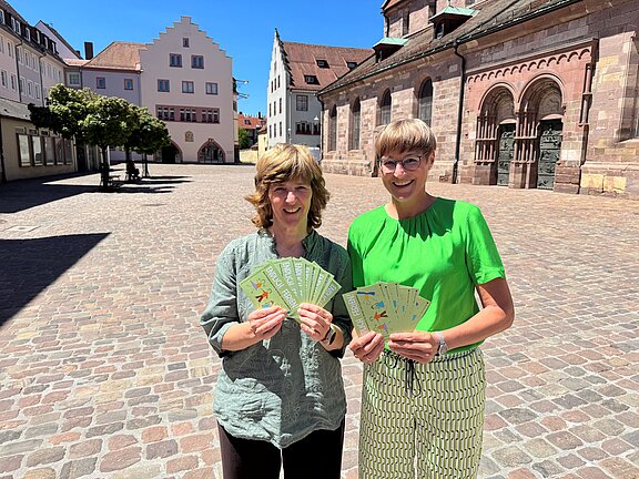 Zwei lächelnde Frauen stehen auf einem gepflasterten Marktplatz und halten jeweils mehrere Informationsflyer fächerförmig in den Händen.
