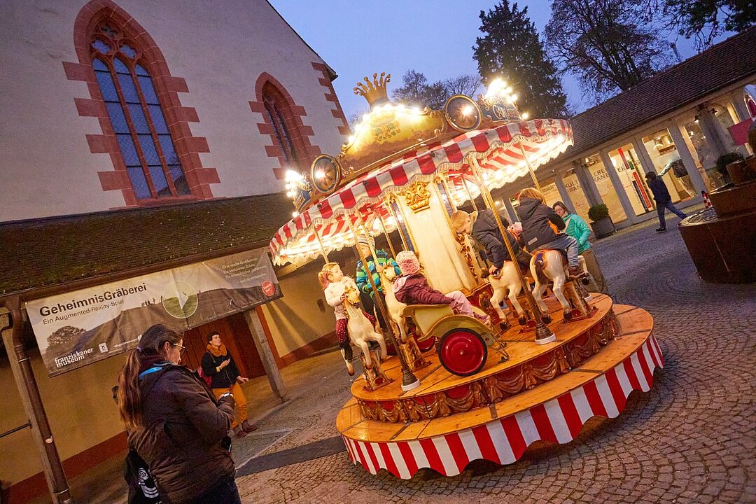 Kinder fahren auf einem kleinen Karussell mit Pferden und einer Kutsche. Im Hintergrund ist ein Gebäude mit großen Fenstern. (KI generiert)